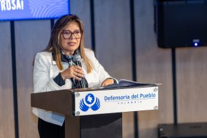 En la imagen aparece la Fiscal Luz Adriana Camargo con una chaqueta blanca en un atril de la defensoria del pueblo. De fondo se ve una pantalla. 