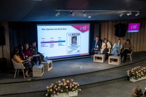 En la imagen aparecen varias personas sentadas en un escenario y una pantalla gigante donde se ve una foto de una niña y colores rosados. 