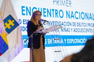 Persona en un escenario institucional interviniendo en un micrófono desde un atril transparente. Al fondo, una pantalla con el texto “PRIMER ENCUENTRO NACIONAL” y referencias a la investigación de delitos contra niños, niñas y adolescentes, así como trata y explotación. A un lado se observa una bandera institucional. La escena corresponde a un evento formal con público.