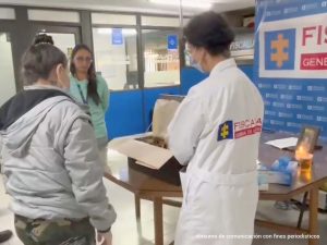 Se observa a una familiar del ciudadano a quien dos funcionarias del laboratorio le muestran los restos de su familiar. Atrás un pendón de la Fiscalía General de la Nación. Adelante una mesa con la Urna funeraria.