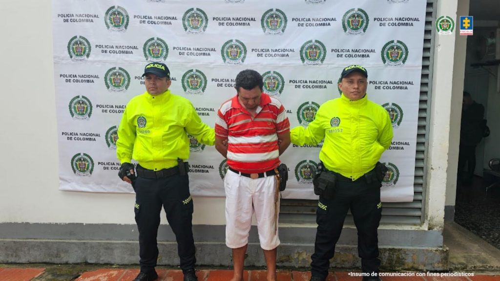 El capturado está esposado con sus manos a la espalda y custodiado por dos uniformados de la policía nacional.