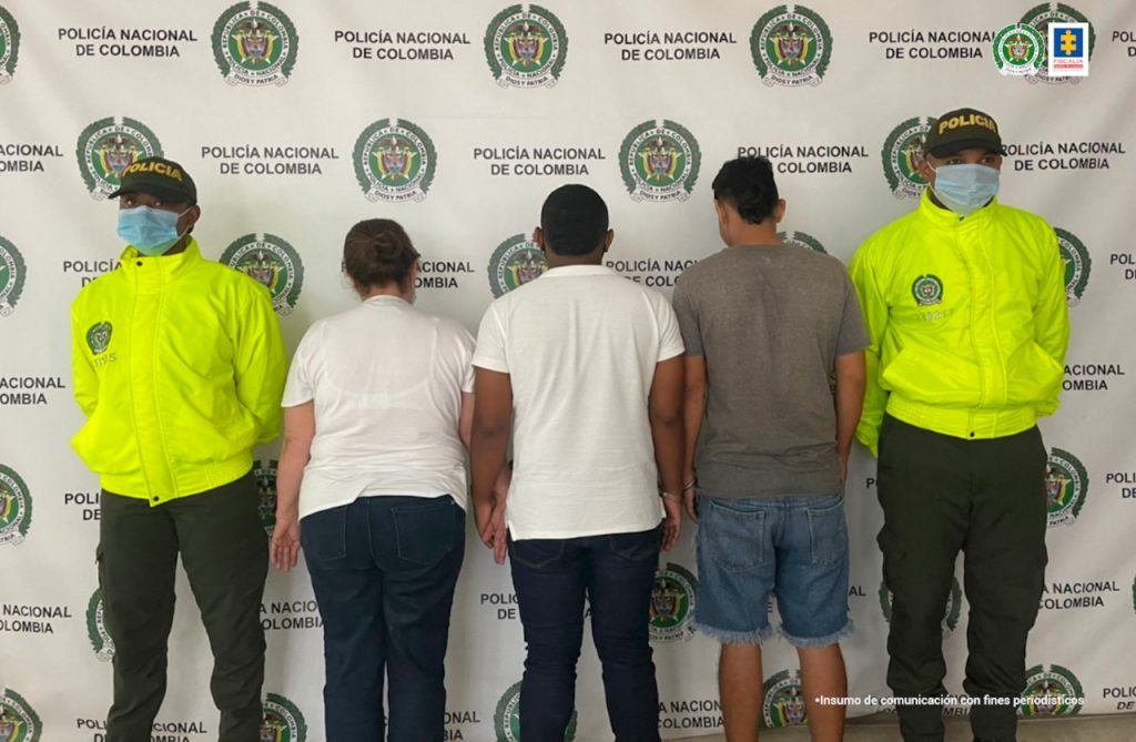 Se ven de espaldas una mujer y dos hombres capturados. A lado y lado personal de la Policía Nacional. detrás se observa un banner de la Policía Nacional