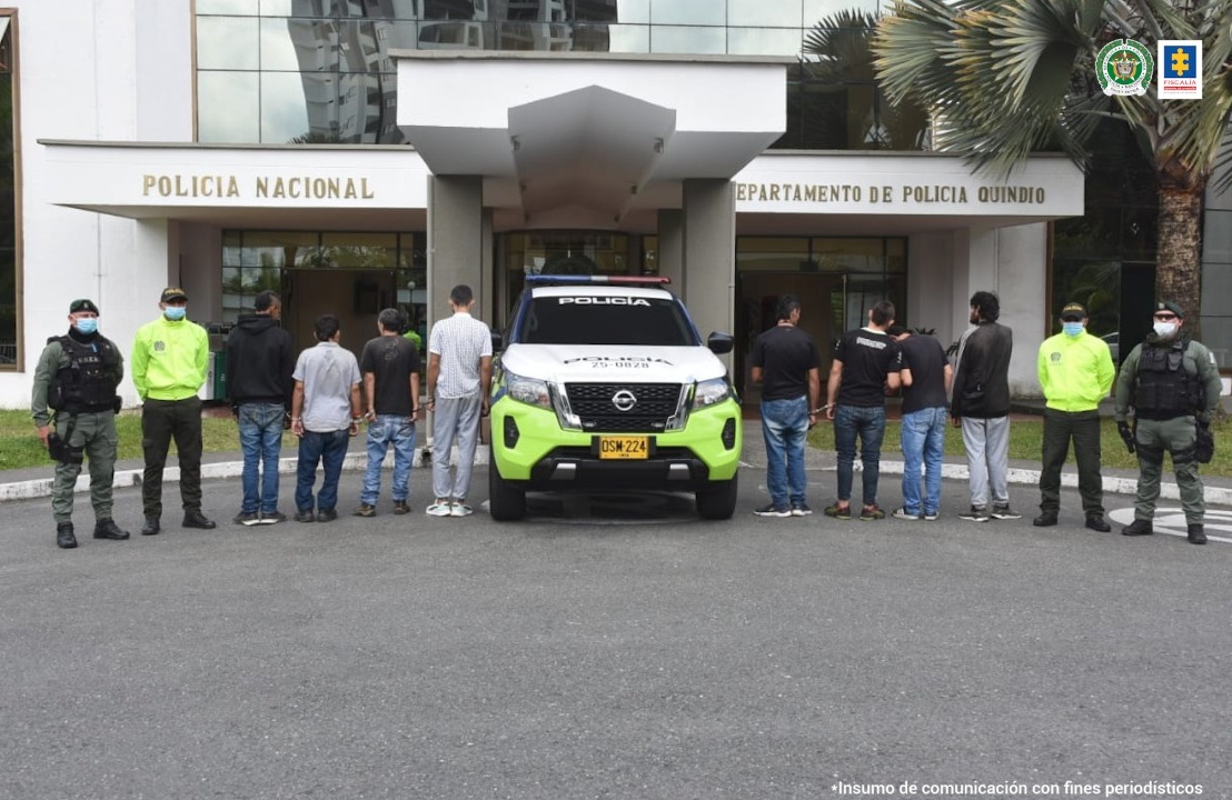 se observan ocho hombres capturados, todos de espaldas formando una fila. En cada lado dos funcionarios de la Policía Nacional. En el centro un vehículo de la Policía Nacional.