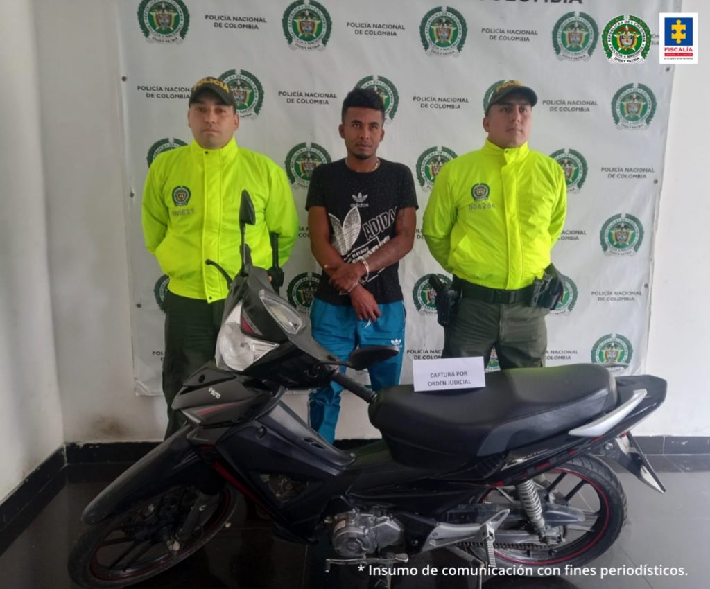 Se observa al capturado en el centro, de frente. En ambos costados un funcionario de la Policía Nacional. Atrás un pendón de la Policía Nacional. Al frente una motocicleta