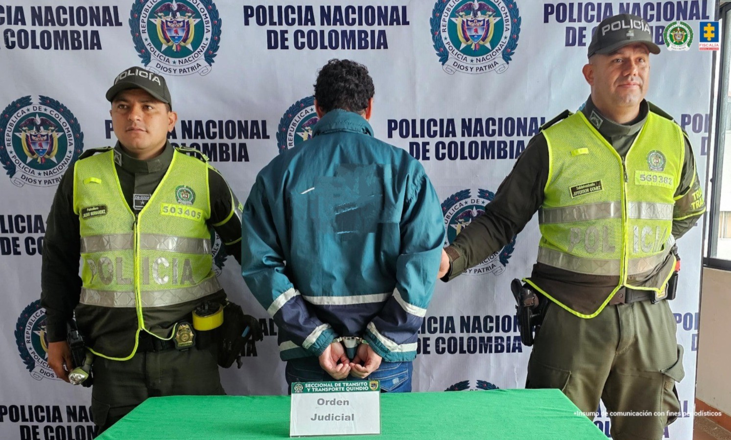 En la fotografía se aprecia de pie, de espaldas a la cámara, con los brazos atrás y esposado al presunto agresor. Viste con chaqueta de color verde con franjas blancas y azules en los puños. A sus extremos hay uniformados de la Policía Nacional. Delante hay una superficie con mantel verde y flequillos de color amarillo y sobre ella hay un letrero que dice: ‘Orden Judicial’. En la parte posterior hay un pendón institucional de la Policía.