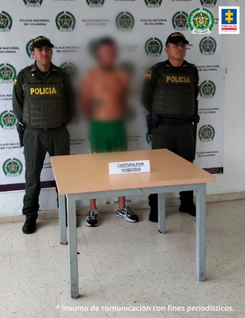 Se observa al detenido de frente. Sobre su rostro un efecto que difumina la imagen. En cada uno de los costados funcionarios de la Policía Nacional. Al fondo un pendón de la Policía Nacional. Al frente del capturado una mesa en donde se ve un cartel con la leyenda: CAPTURA POR HOMICIDIO