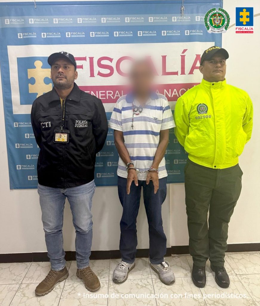 capturado de frente custodiado por investigador del CTI de la Fiscalía y un policía. Detrás banner de la Fiscalía.