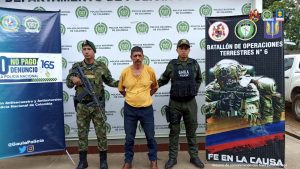 Eduardo Antonio Mejía Ortiz es imputado por extorsión agravada. Texto Alternativo: capturado es custodiado por un policía y un militar. Banner de Policía y Ejército Nacional.