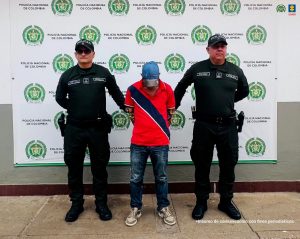 capturado de frente con gorra custodiado por policías. Detrás banner de la Policía Nacional.
