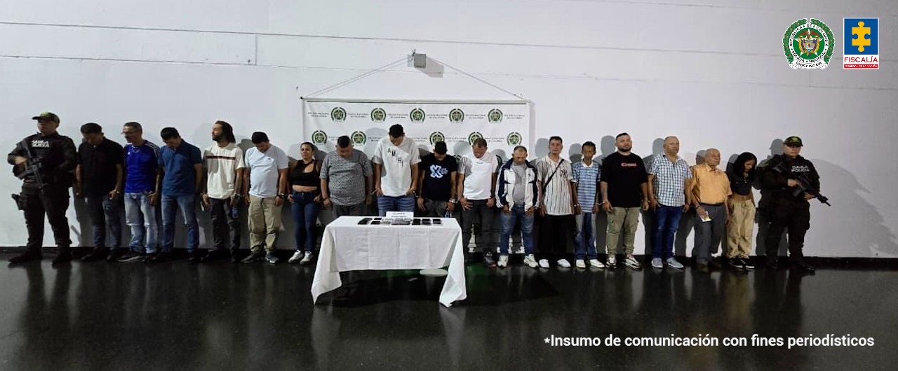 Un grupo de 21 personas, hombres y mujeres, permanece en fila con las manos esposadas frente a una pared blanca. Detrás de ellos hay un pendón con los logos de la Policía Nacional, y a los costados se observan dos uniformados armados de la Policía. En el centro, sobre una mesa cubierta con un mantel blanco, reposan varios celulares incautados.