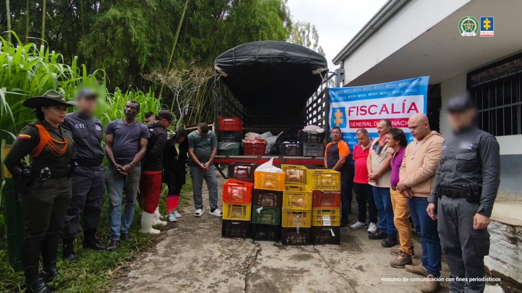 Se observan los capturados alrededor de canastillas que contienen material incautado, Atrás un camión en el que se observan más canastillas y a un costado un pendón de la Fiscalía General de la Nación.  Los custodian dos funcionarios del Cuerpo Técnico de Investigación CTI y uno de Policía Nacional. Atrás, un pendón de la Fiscalía General de la Nación