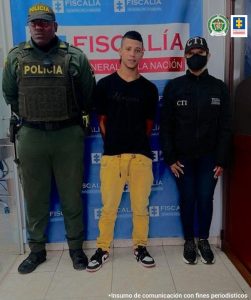 En la foto aparece Jaider Suárez Bedoya con los brazos atrás; vestido con camiseta negra, pantalón de color mostaza y tenis negro con blanco. Al lado derecho de él hay un uniformado de la Policía Nacional y al lado izquierdo un servidor del CTI de la Fiscalía. Detrás de ellos aparece un pendón institucional de la Fiscalía.