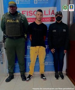 En la foto aparece Jaider Suárez Bedoya con los brazos atrás; vestido con camiseta negra, pantalón de color mostaza y tenis negro con blanco. Al lado derecho de él hay un uniformado de la Policía Nacional y al lado izquierdo un servidor del CTI de la Fiscalía. Detrás de ellos aparece un pendón institucional de la Fiscalía. 