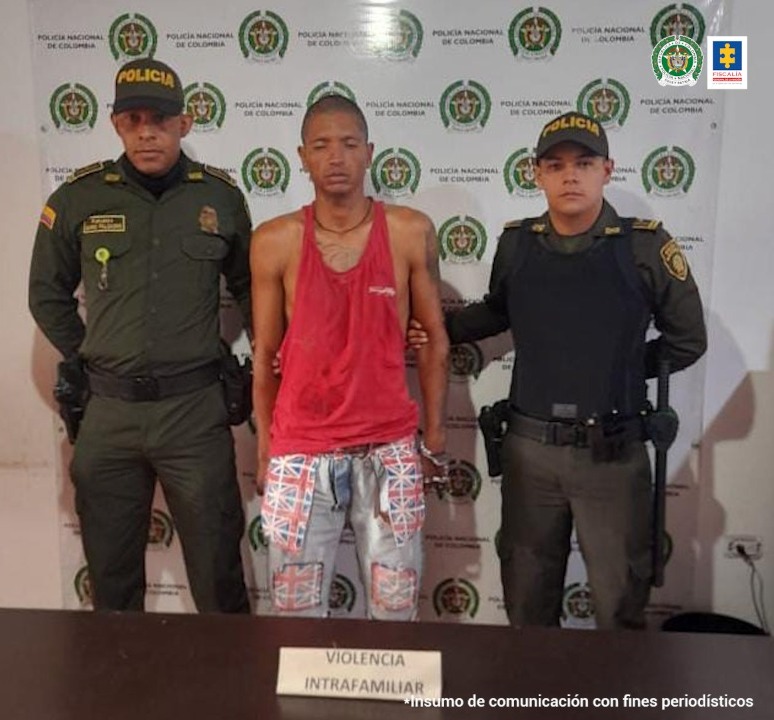 En la foto aparece de pie Jhon Harold Trujillo López, vestido con camiseta esqueleto roja y pantalón claro con estampados. A lado y lado de él hay dos uniformados de la Policía Nacional. Delante de ellos hay una mesa con un letrero que dice ‘violencia intrafamiliar’. Detrás de ellos hay un pendón institucional de la Policía.