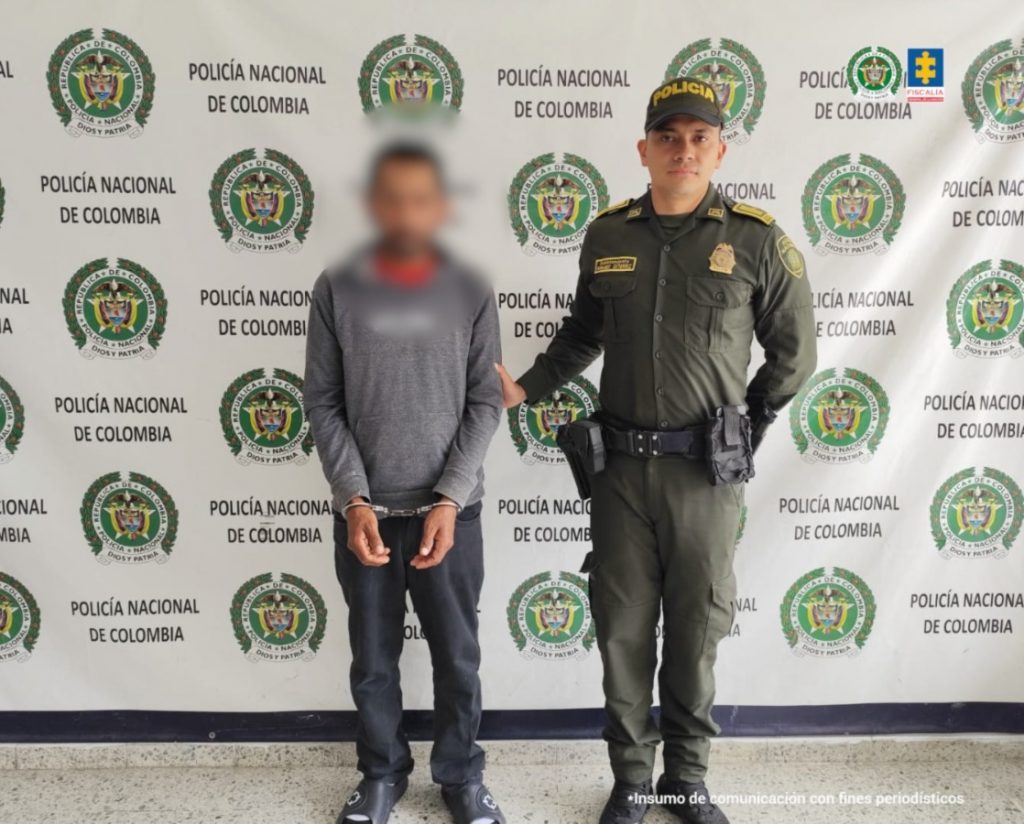fotografia en donde se ve al capturado esposado custodiado por un policia, detras de ellos hay un baking de la policia