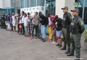 Las fotografías están tomadas en diagonal de derecha a izquierda y viceversa. Se aprecian los presuntos implicados en hilera y esposados entre sí. Hay 4 mujeres y 13 hombres. A sus extremos hay uniformados de la Policía Nacional. Detrás de ellos hay un pendón institucional de la Policía.
