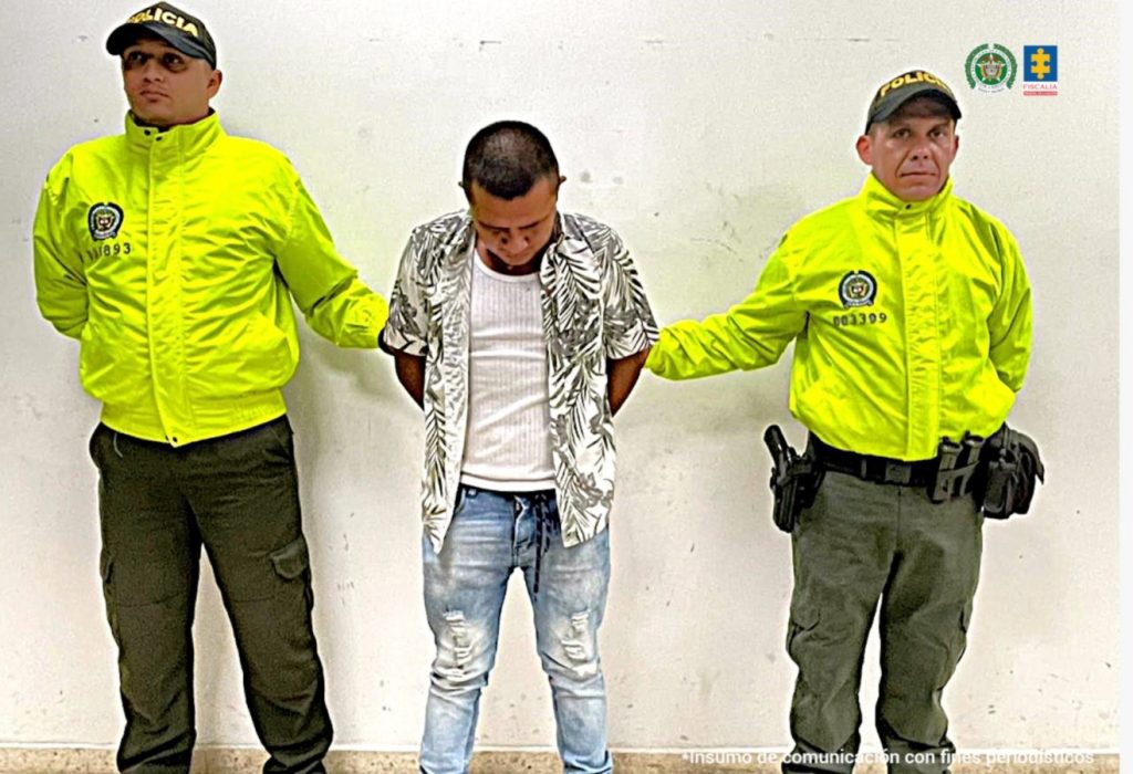 El capturado está custodiado por dos uniformados de la policía nacional, esposado con sus manos en la espalda.
