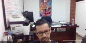 Hombre de mediana edad, con gafas y cabello corto, observado desde arriba mientras está sentado en una oficina. Al fondo se ven un escritorio, equipos de oficina, una impresora, documentos y un tablero con anotaciones, en un ambiente de trabajo cerrado.