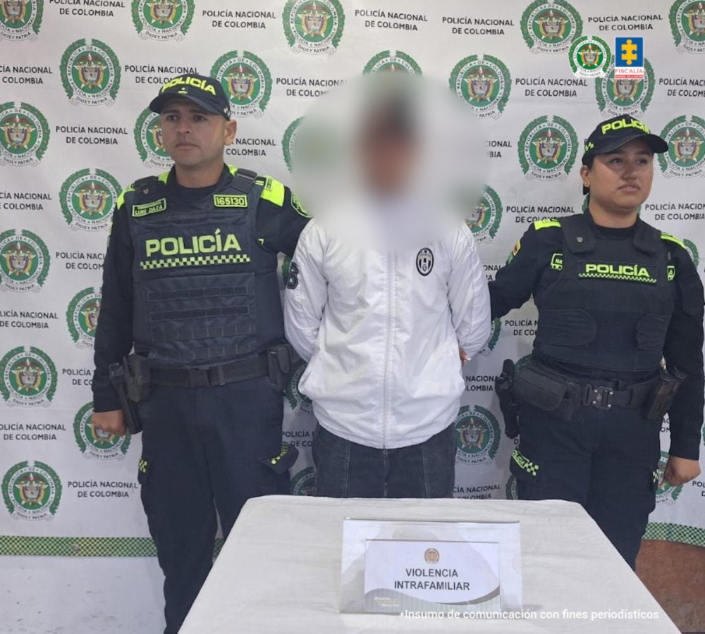 fotografia en donde se puede ver al capturado con chaqueta blanca en medio de dos policias, en frente de ellos hay una mesa con elementos incautados y detras de ellos hay un baking de la policia