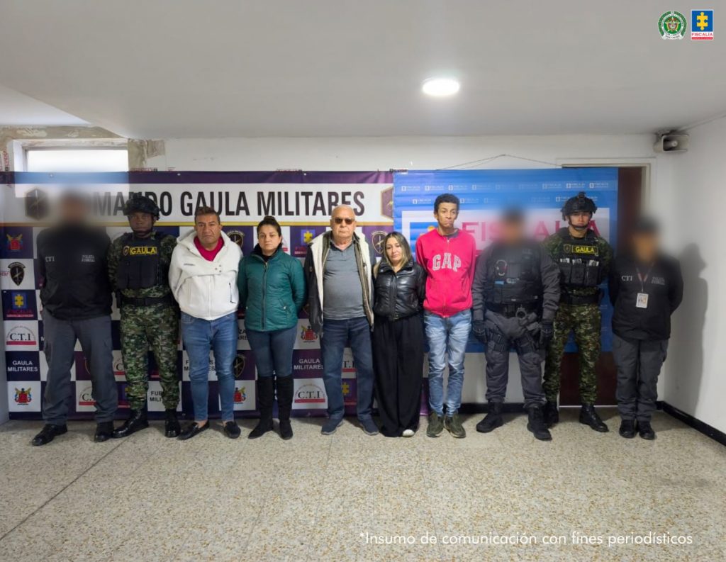 Imagen de un grupo de once personas en una sala institucional. En el centro aparecen seis personas capturadas, hombres y mujeres, de pie y alineadas frente a un fondo con logos de la Fiscalía General de la Nación, el CTI y el Gaula Militar. A los lados se observan uniformados del Gaula y del CTI con prendas tácticas. La fotografía corresponde a un procedimiento judicial relacionado con delitos fiscales y lavado de activos.