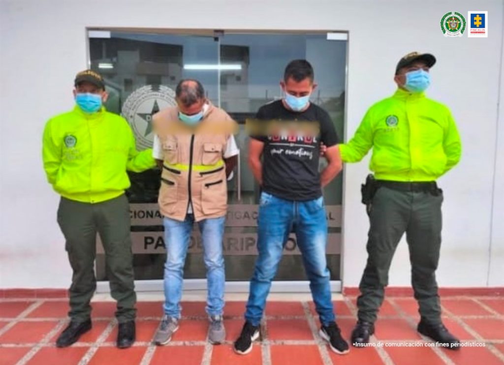 Los dos capturados están custodiados por dos policías, sus manos están esposadas a su espalda, y tienen tapabocas.