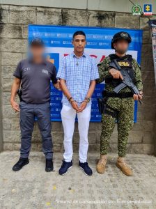 En la fotografía, está un hombre joven, trigueño, delgado, de pie, con los brazos adelante esposados, vestido con pantalón blanco, camisa de cuadros azul con blanco y tenis azul oscuro, custodiado por un uniformado del CTI y otro de la Policía Nacional. Detrás de ellos hay un pendón de la Fiscalía.