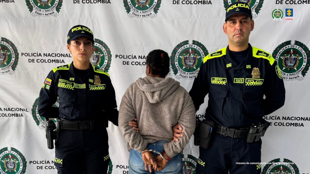 En la imagen se ve a tres personas de pie frente a un fondo blanco con logos repetidos de la Policía Nacional de Colombia. A los extremos izquierdo y derecho hay dos uniformados de la Policía, una mujer y un hombre, con uniforme oscuro y detalles reflectivos en amarillo. Están serios y de frente a la cámara. En el centro, de espaldas, está una mujer con las manos esposadas detrás de la espalda. Lleva una chaqueta gruesa de color gris claro con capucha y jeans azules. Su cabello está recogido en trenzas.