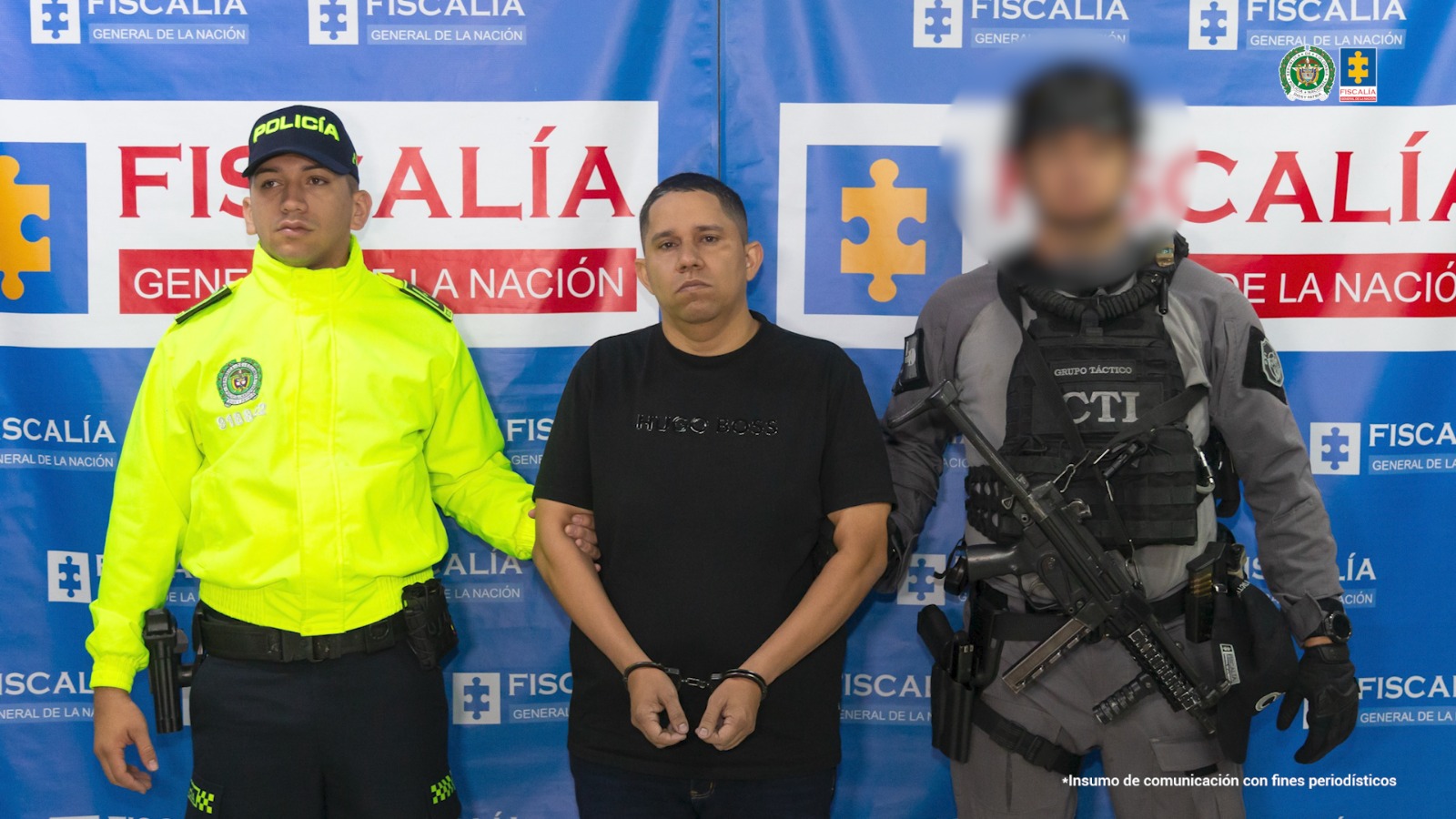En la imagen aparecen tres hombres frente a un fondo azul con logotipos de la Fiscalía General de la Nación. En el centro está un hombre vestido de camiseta negra, esposado de las manos, con expresión seria. A su lado izquierdo hay un policía con chaqueta reflectiva amarilla y gorra negra con la palabra “Policía”. A la derecha se ve un integrante del CTI con uniforme táctico gris, chaleco antibalas y un arma larga.