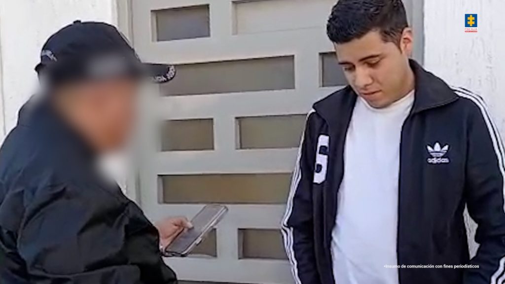 En la imagen se observa a dos hombres de pie frente a una puerta de color claro. A la izquierda, un funcionario del CTI, con gorra y chaqueta oscura, sostiene una libreta electrónica mientras realiza un procedimiento. Su rostro aparece desenfocado. A la derecha, un joven de cabello corto, con chaqueta deportiva negra con franjas blancas y camiseta blanca, baja la mirada con expresión seria. En la parte superior derecha se ve el logotipo de la Fiscalía General de la Nación.