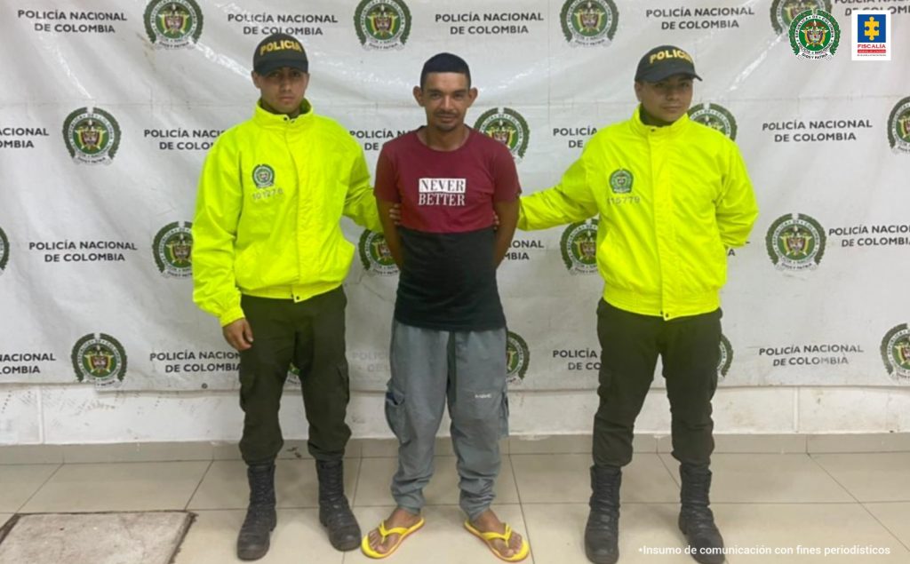 El capturado esta con sus manos a la espalda, esposado y custodiado por dos uniformados de la Policía Nacional y detrás de ellos está un pendón institucional