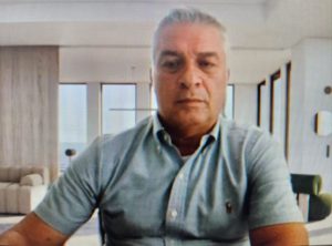 Un hombre de cabello canoso y corto, con camisa de manga corta color azul claro, aparece sentado frente a la cámara en un espacio luminoso. El fondo muestra una sala amplia con sofás claros, ventanales grandes y cortinas que dejan entrar la luz natural. Su expresión es seria y mira directamente hacia el frente.