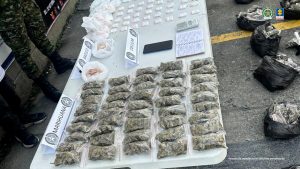  Sobre una mesa se observan varias bolsas con material vegetal y un aviso que dice MARIHUANA. Además, otras bolsas con un aviso que dice BASUZO. Un celular con el aviso CELULAR, además de un cuaderno y una gramera. Al lado, una serie de bolsas pequeñas. En el piso, al frente de la mesa, se ven algunas bolsas con material vegetal