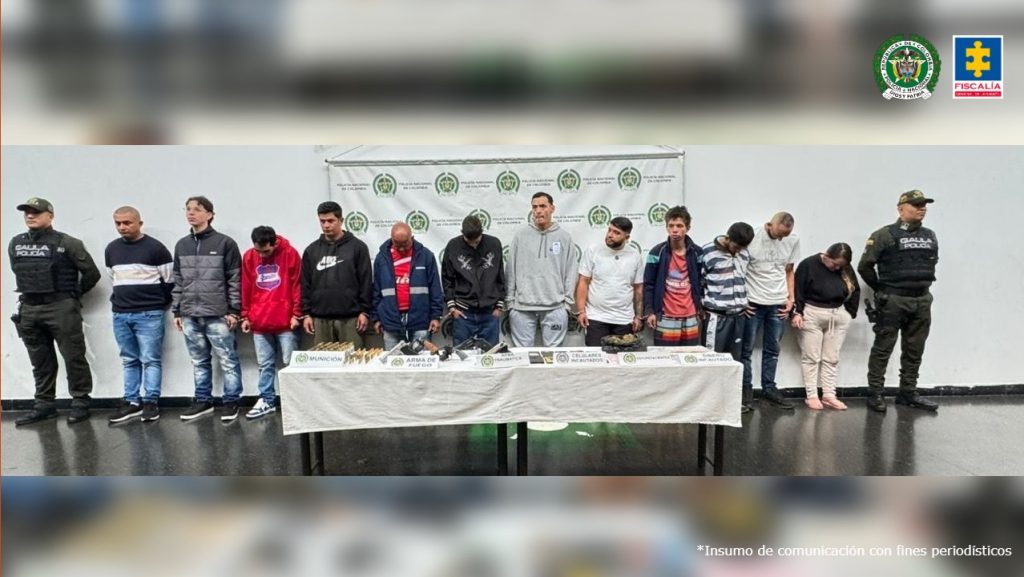 En la imagen se observa una fila de doce personas detenidas, escoltadas por dos uniformados del Gaula de la Policía Nacional ubicados a los extremos. Los capturados están de pie frente a una mesa en la que se exhiben armas de fuego, munición, teléfonos celulares y otros elementos incautados. Al fondo se ve un telón blanco con los logotipos de la Policía Nacional. En la esquina superior derecha aparecen los escudos de la Fiscalía General de la Nación y de la Policía Nacional.