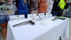 En una mesa aparecen los elementos incautados como armas, celulares estupefacientes y de fondo, están los capturados esposados y custodiados por uniformados de la Policía Nacional.