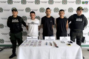Los capturados aparecen esposados, y frente a ellos en una mesa están elementos incautados como dinero en efectivo, armas blancas, equipos celulares. Están custodiados por hombres del Gaula Policía.