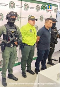 Los capturados están con sus manos esposadas a la espalda, y custodiados por uniformados de la Policía, en una mesa frente a ellos, está el arma de fuego incautada y equipos celulares.
