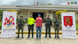 El procesado se encuentra esposado de espaldas y en custodia por uniformados de la Policía y Ejército Nacional. Al lado hay dos backing del Ejército y Batallón de Infantería No. 25.