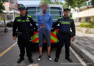 En la fotografía se aprecia de pie y con los brazos atrás al imputado. Viste un cobertor azul, bermuda azul y zapatos negros con blanco. Se encuentran en una vía pública. A sus extremos hay uniformados de la Policía Nacional. Detrás de ellos hay un vehículo.