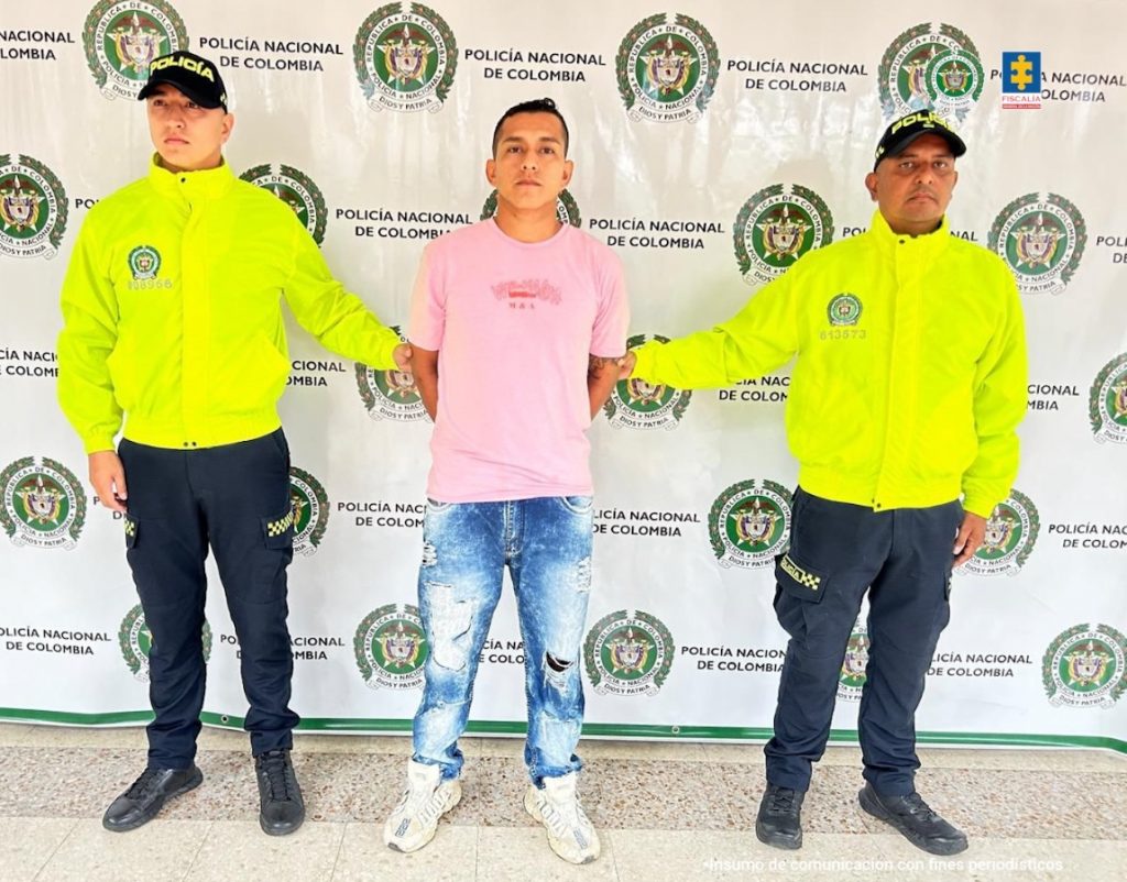 El imputado se encuentra en custodia por dos uniformados de la Sijin de la Policía Nacional. Detrás hay un backing de la Policía