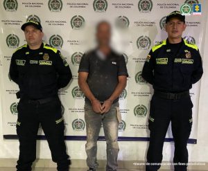 En la foto se observa al presunto agresor de frente y su rostro se ve difuminado por un efecto de blurr que lo cubre. A cada lado lo custodia un funcionario de la Policía Nacional. Atrás, un pendón de la Policía Nacional.