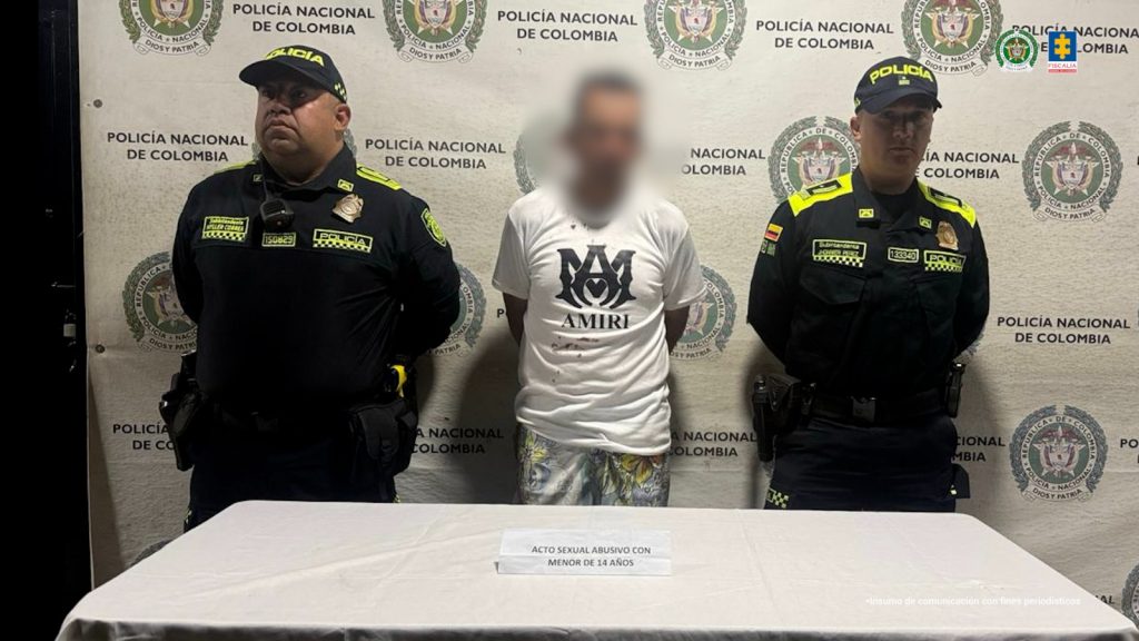 Se obseerva al capturado de frente con las manos atrás. A cada lado lo custodia un funcionario de Policía Naciones. Atrás un pendón de Policía nacional. Adelante, una mesa sobre la cual un cartel que dice : ACTO SEXUAL ABUSIVO CON MENOR DE 14 AÑOS