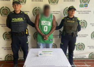 Se observa al capturado de frente, usa camiseta verde y pantalón azul. A cada lado lo custodia un funcionario de la Policía Nacional. Atrás un pendón de Policía Nacional. Adelante una mesa con el aviso que dice Orden Judicial..