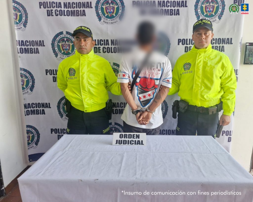 Se observa al capturado, de espaldas, con un efecto de difuminado sobre su cabeza. A cada costado lo custodia un funcionario de Policía nacional. Atrás, un pendón de Policía Nacional. Adelante, una mesa con un cartel que dice: ORDEN JUDICIAL.