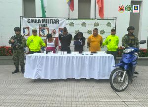 En la fotografía se encuentran los dos hombres y las dos mujeres capturadas, con un servidor de la Policía y otro del Ejército Nacional a cada lado, con una mesa en la que están las armas de fuego y los elementos incaurados. 