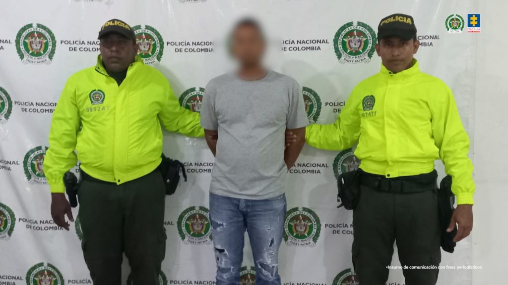 En la fotografía se aprecia de pie y con los brazos atrás al presunto implicado en el abuso sexual a su hijastro. Viste con camiseta de manga corta y de color gris; jean azul desgastado y zapatillas de color negro con zuela blanca. A sus extremos hay uniformados de la Policía Nacional. Detrás de ellos hay un pendón institucional de la Policía.