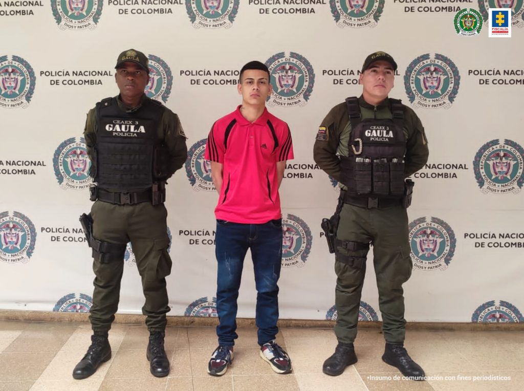 AL CENTRO SE OBSERVA UN CAPTURADO CON CAMISETA ROJA . A CADA COSTADO UNIFORMADOS DE POLICIA NACIONAL Y AL FONDO UN PENDÓN DE POLICÍA NACIONAL