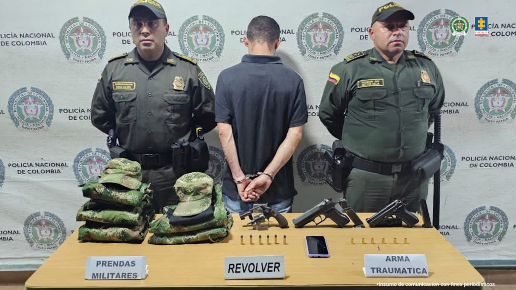 Se observa al capturado de espaldas, esposado.  A cada costado un funcionario de Policía Nacional, atrás un pendón de Policía Nacional y adelante una mesa con el material incautadoPIE DE FOTO: Andrés Felipe Agudelo Orozco fue capturado en La Virginia. Al parecer portaba armas, munición y prendas  de uso de las fuerzas armadas.