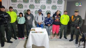Se observan CINCO personas en fila y de frente, A cada costado funcionarios de Policía Nacional, en uno de los costados un canino de la Policía Nacional. Atrás un pendón de Policía NAcional, al frente una mesa con elementos incautados