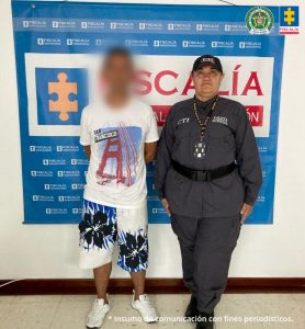 En la fotografía aparece de pie y los brazos atrás Albdul García González. Viste camiseta blanca, de manga corta, con estampado de colores azul y rojo; pantaloneta blanca con estampado azul y negro; así como zapatillas blanca. Al lado izquierdo de él hay una servidora del CTI. Detrás de ellos hay un pendón institucional de la Fiscalía.