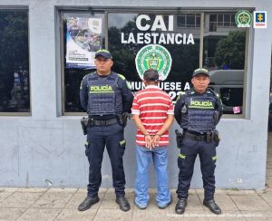 Se visualiza al capturado de espaldas junto a dos uniformados de la Policía Nacional. Detrás se observa logo que identifica a una estación de Policía